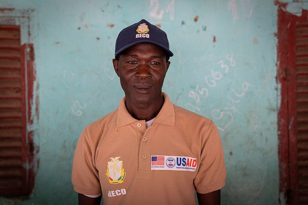 A Guinean man wearing a baseball cap and a polo shirt with the USAID logo on it.