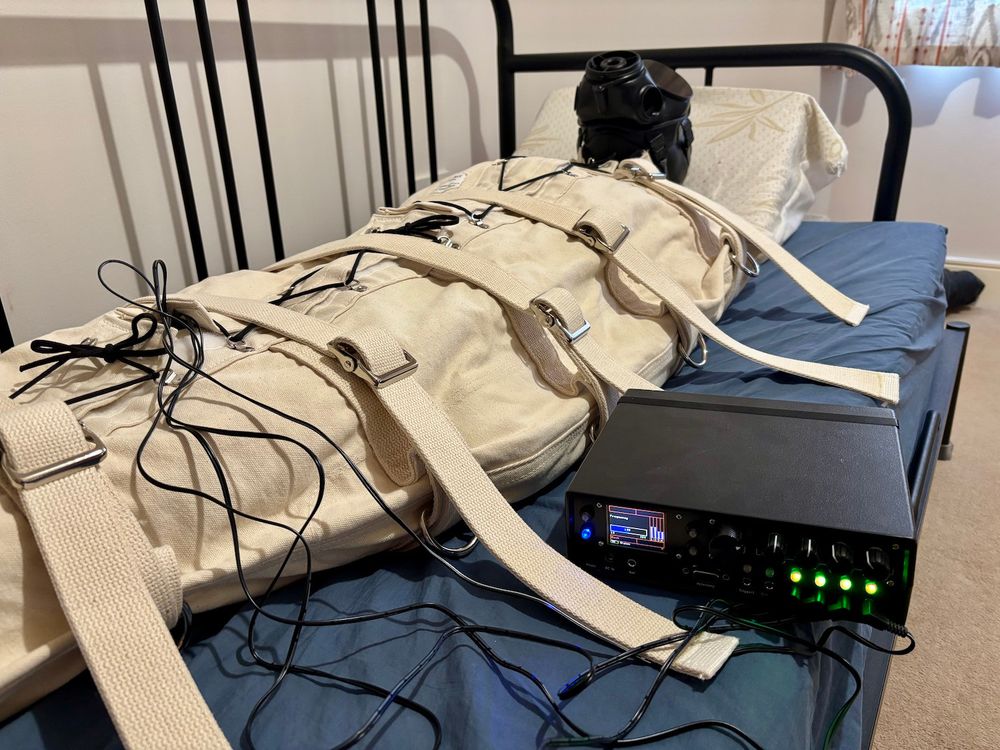 Person bound in a white fabric sleepsack. They are wearing a gas-mask. Estim device with several cables coming from it is laying next to the sleepsack.