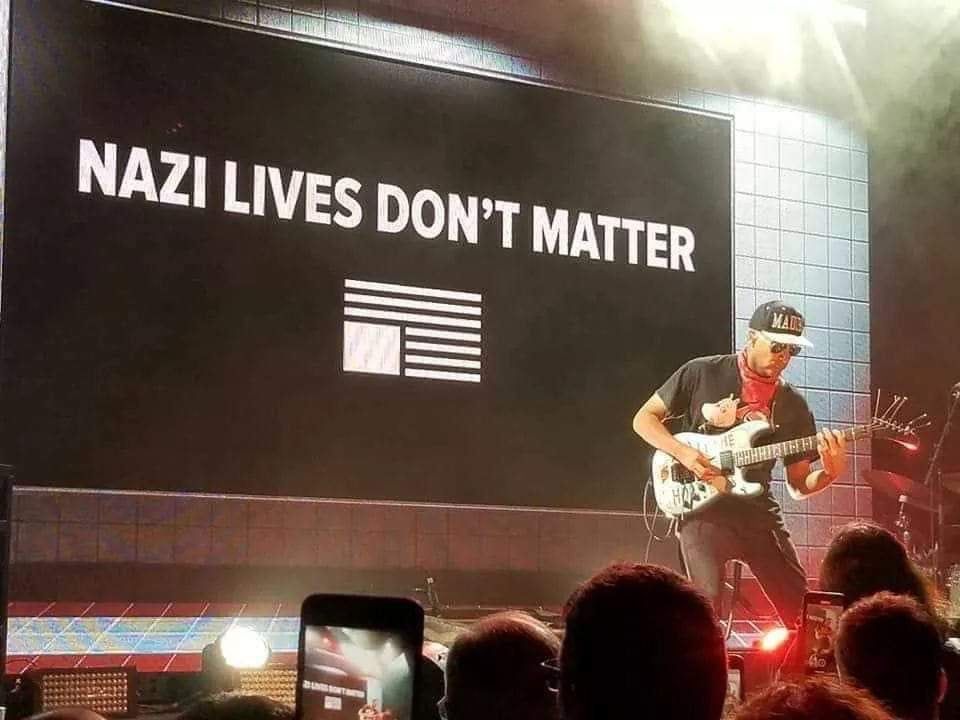 Tom Morello playing guitar with “Nazi Lives Don’t Matter” sign behind him.