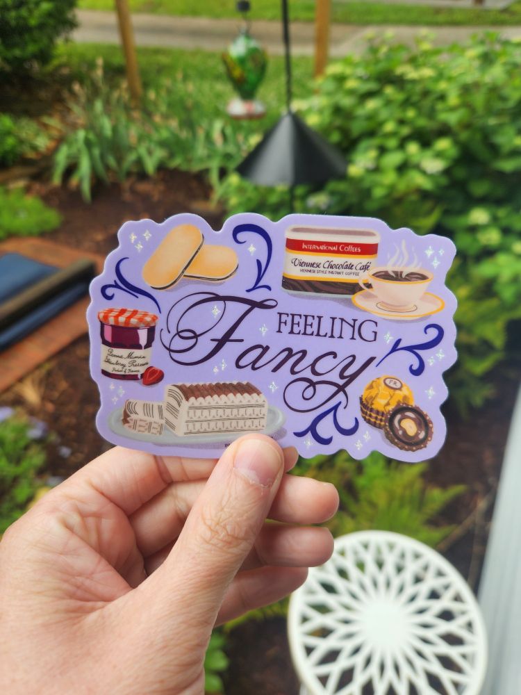 This sticker says "feeling fancy" in script font on a periwinkle background, and is surrounded by fancy little treats like Milano cookies, Ferraro rocher chocolates, and Bonne Maman strawberry preserves.
