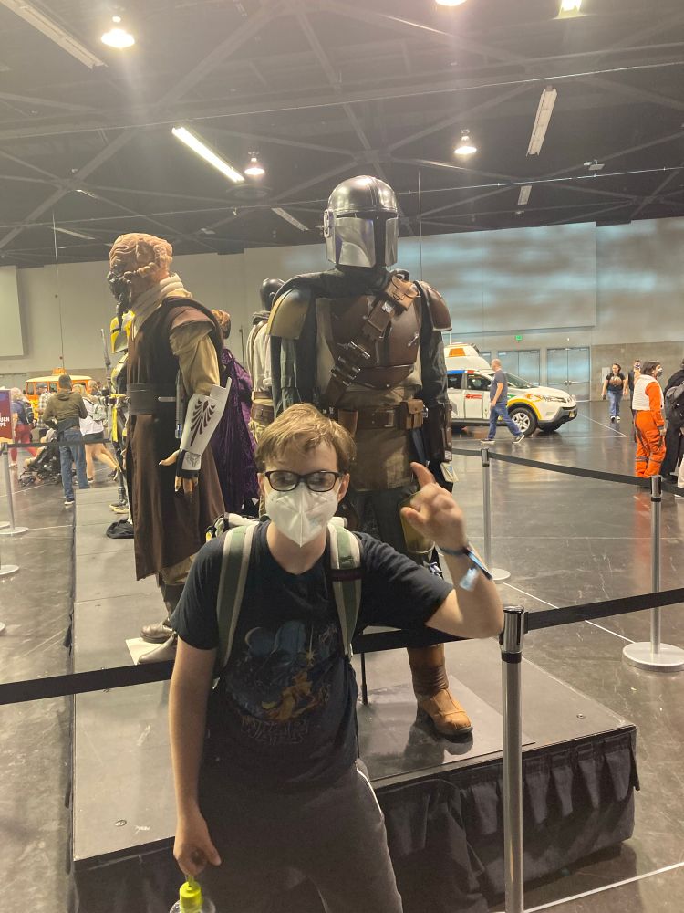 Eli posing in an A NEW HOPE T-shirt in front of a Din Djarin costume from THE MANDALORIAN at an exhibition at Star Wras Celebration Anaheim 2022.