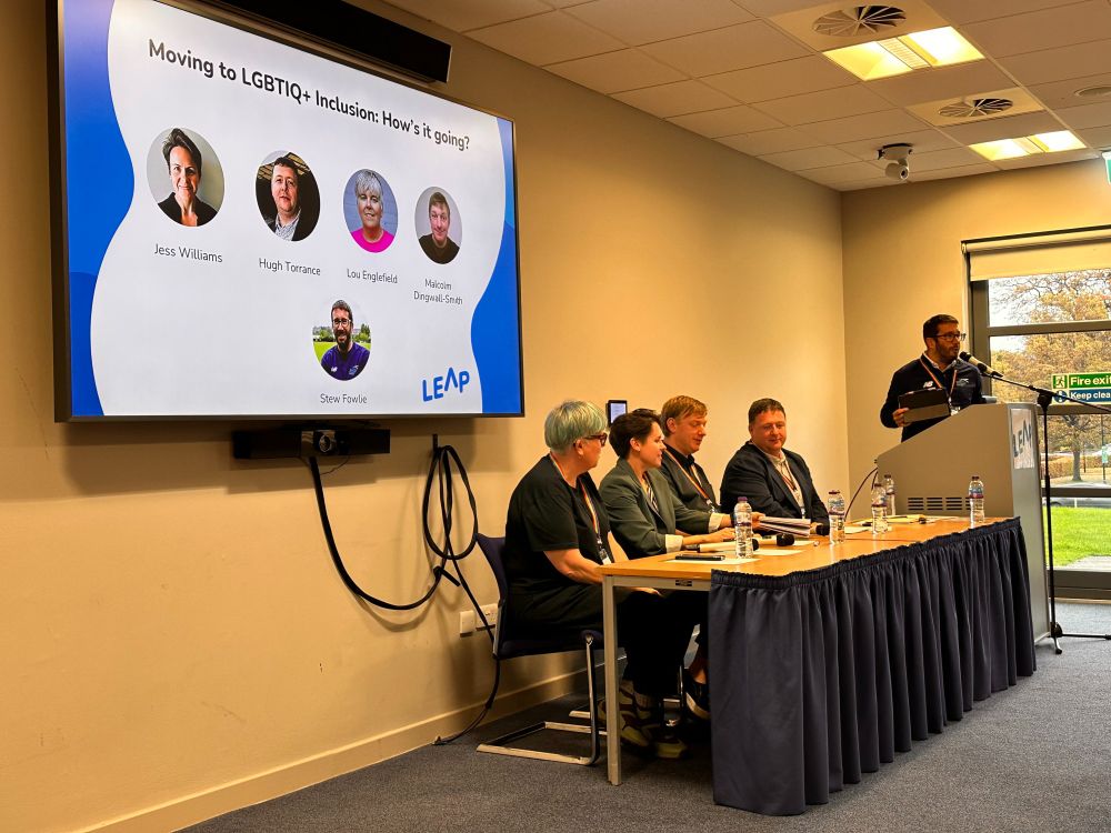 Four people sitting at a top table of a conference room ready to speak as part of a panel. Behind them is a screen that reads "Moving to LGBTIQ+ Inclusion: How is it going?" The leader of the panel, Stew, is standing on the right of the top table. 