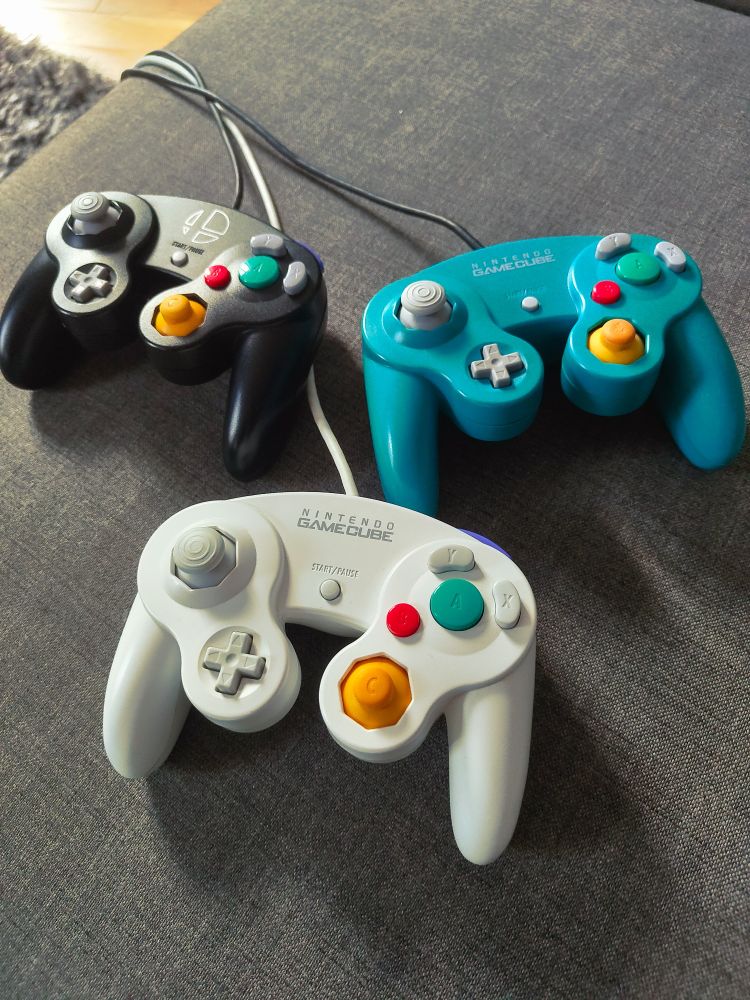 GameCube controllers (a SSBU black one, a JP White one, and an electric blue one) on a gray couch