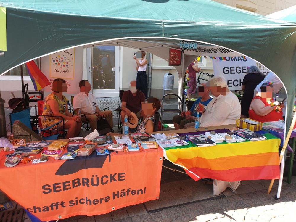 Foto eines bunten Infostandes von verschiedenen Akteur*innen von der Seebrücke Brühl, Gemeinsam für Brühl, der Brühler Initiative für Völkerverständigung, den Omas gegen Rechts REK, dem Fahnenprojekt "Kein Ort für Rassismus" und a.d.a.

Im Bild sieht man einen oliv-grünen Pavillon,  unter dem sich 8 sommerlich gekleidete Menschen aus den verschiedenen Initiativen & Organisationen befinden. Auf Tischen befinden sich viele Flyer, Broschüren & Giveaways. Vorne links hängt eine "Seebrücke - schafft sichere Häfen!" Fahne, rechts eine Regenbogenfahne. Im Hintergrund hängt weitere Regenbogen-Deko, eine Fahne der Omas gegen Rechts, ein Banner von Gemeinsam für Brühl & von der BIfV, sowie ein kleines Campact-Pkakat, auf dem "Rote Karte für die AfD" steht.

Ganz im Hintergrund telefoniert eine weitere Person. Die Gesichter aller Menschen wurden verpixelt.