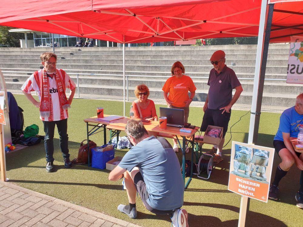 Ein Läufer bindet sich vor dem Spenden-Stand die Schuhe.

Zu sehen sind neben dem Herrn, der der Kamera den Rücken zudreht, mehrere Personen von der Seebrücke Brühl & Helfer*innen, die hinter & neben dem Stand unter einem roten Pavillon stehen und sitzen.

Unten rechts sieht man ein Demo-Plakat, welches am Pavillon befestigt ist. Darauf steht "Sicherer Hafen Brühl". Darüber ein Bild der Brunnen-Figuren des Max-Ernst-Brunnens, ein Wahrzeichen Brühls.