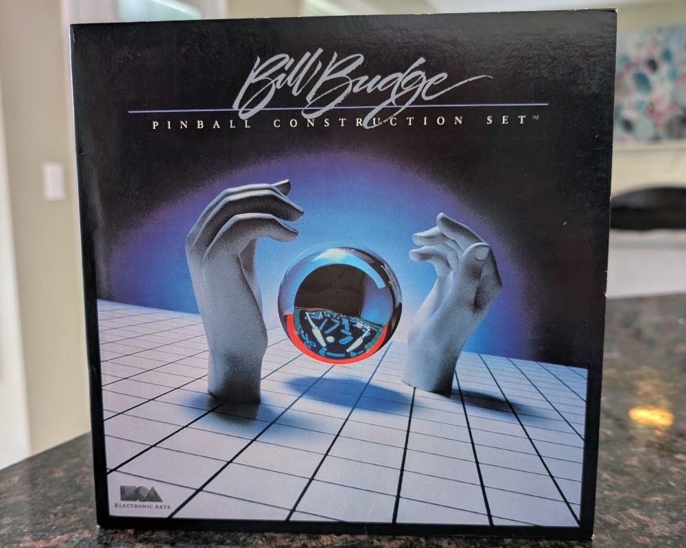 The front cover of Pinball Construction Set (with designer Bill Budge's name prominently features). It has a retrowave aesthetic with a silver ball floating between two hands. They're place on a seemingly infinite grid.