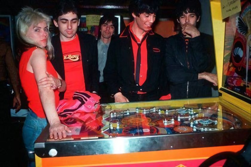 Debbie Harry is seen playing pinball with the other members of Blondie, wearing a red top and blue jeans, leaning casually on the pinball machine, which has a bright yellow and red design. The group appears relaxed and engaged.