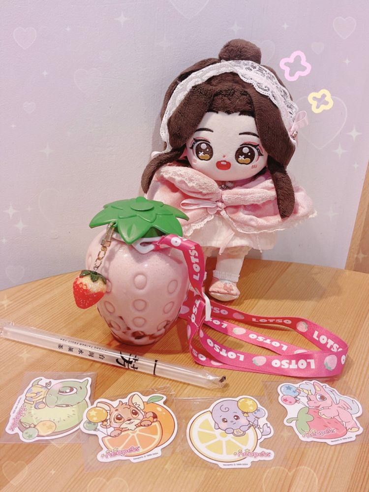 a xie lian minidoll stands next to a strawberry shaped cup filled with strawberry milk tea with a charm and strawberry themed lanyard. there’s also free neopets stickers set up below