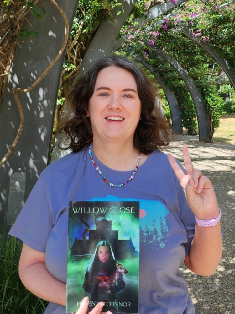 Me, in a blue tshirt with a arch of flowers background, holding a copy of my novella Willow Close, and doing this: ✌️