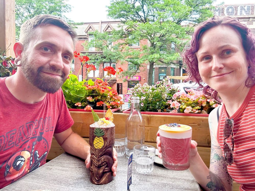 A man and a woman sitting on a restaurant patio, both with tiki drinks in their hands.