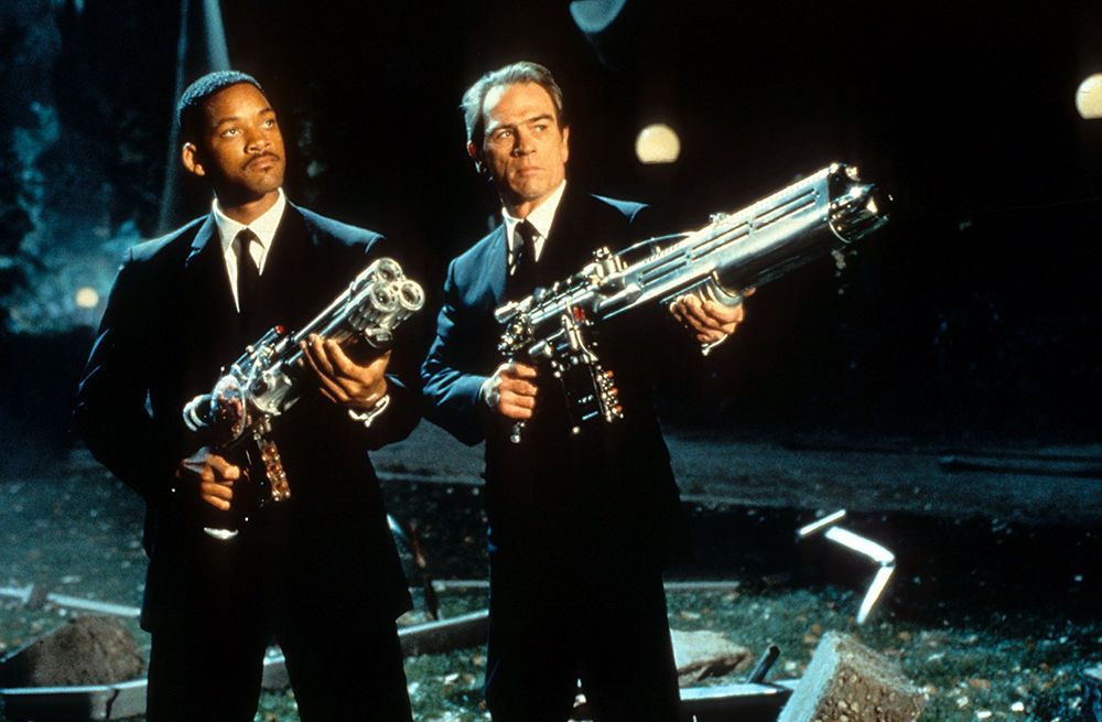 Two men, portrayed by Will Smith and Tommy Lee Jones left to right, hold up chrome sci-fi guns at a threat off camera. They stand in a location covered in rubble and greenery.