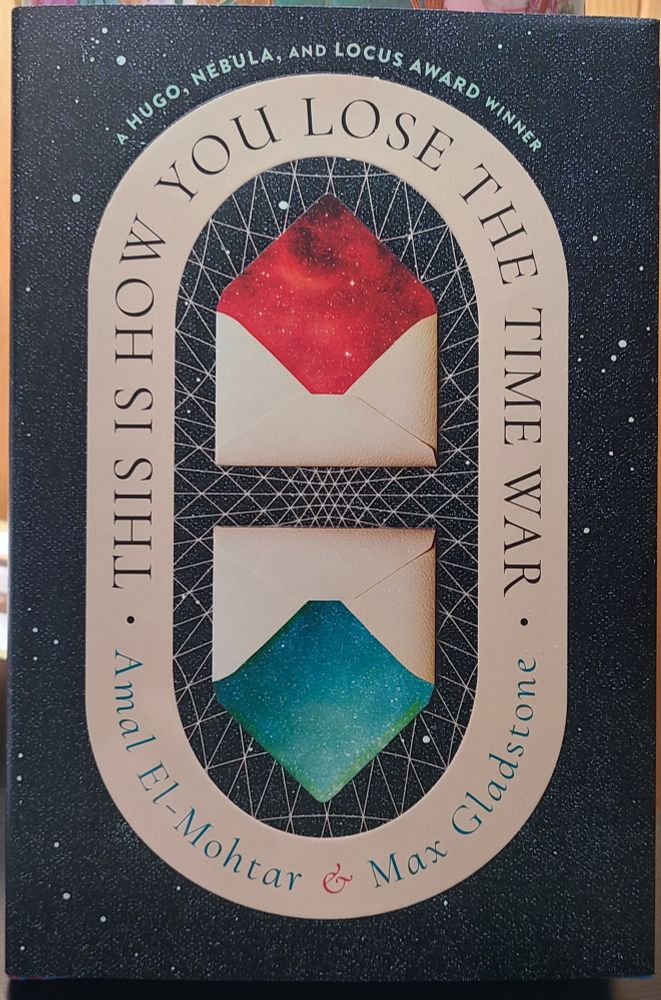 The dust jacket of the new hardcover edition of THIS IS HOW YOU LOSE THE TIME WAR, by Amal El-Mohtar & Max Gladstone, which has a pleasing design over a starfield, with the title & authors' names in an ivory colored oval, inside of which are two envelopes with the flaps open, one with red spot gloss envelope liner & the other upside down below the first with blue spot gloss.