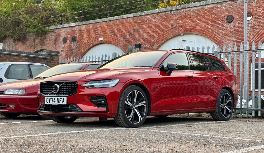 A red 2024 Volvo V60 estate parked in front of a set of old brick arches and a metal fence on an industrial estate. In the background is a Citroen Xantia.