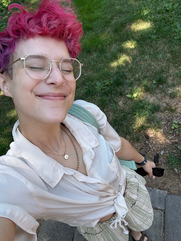 grace selfie from a high angle. their eyes are squinted closed behind gold wire framed glasses. it has magenta and purple short hair. wearing gold jewelry (nose hoop, taurus necklace, gold rope chain), white short sleeve button down tied at the waist, green and cream striped shorts, green bag strap over one shoulder. grass in background