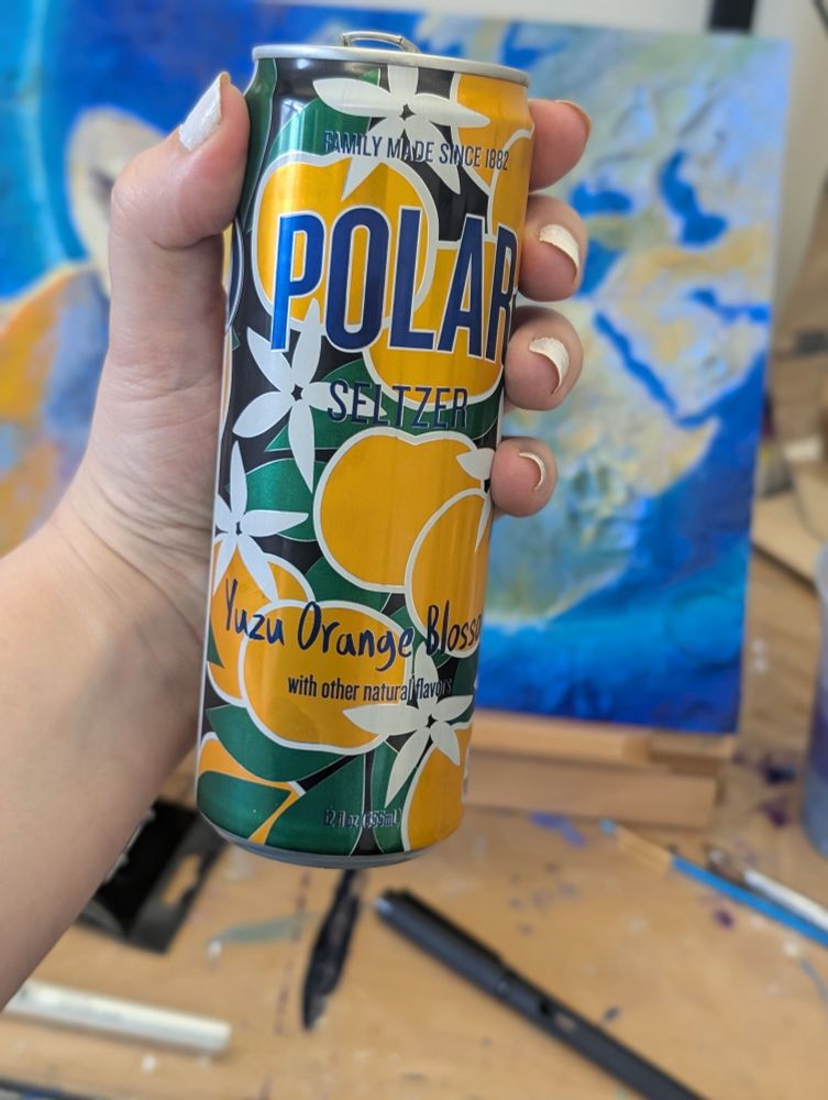 A feminine hand with white nails holds a can of Yuzu Orange Blossom seltzer by Polar Springs with a blurry painting and artist tools in the background. 