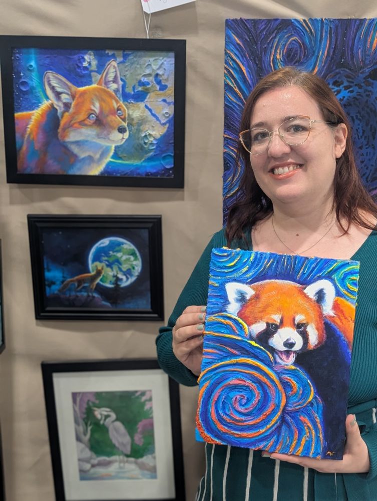 An artist holding a painting of a red panda standing in front of a group if paintings, top left of a fox head with an earth in the background, middle of a full body fox with earth, and the bottom of a great blue heron. 