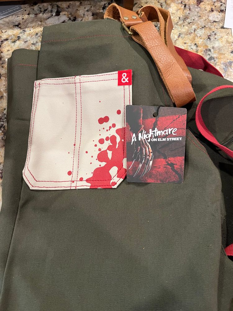 The top front pocket (white with blood splatters) of an olive green apron with a tan leather neck strap, folded and resting on a brown granite kitchen counter, and the tag from the apron showing it is A Nightmare on Elm Street collaboration 