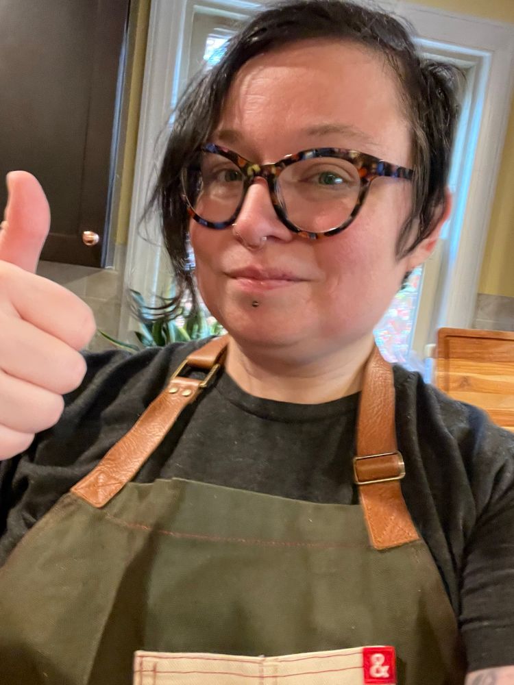 A very normal selfie of a very normal woman with short hair and cat-eye tortoiseshell glasses in her kitchen, with a cutting board in a drying rack behind her and a plant peeking out next to a window, giving a dorky thumbs-up about the awesome apron she’s wearing. (The woman is me.)