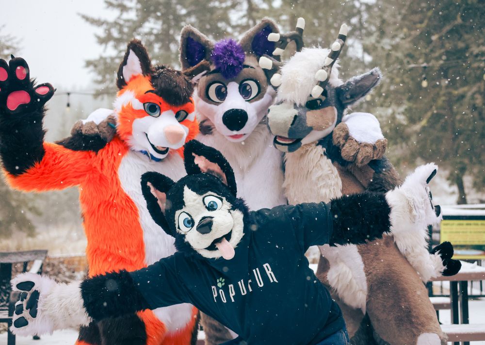 Buddy, the black and white husky wearing a black t-shirt and blue denim jeans, poses low with his arms stretched out while in front of his buddies standing behind him - Kyred the orange and black fox-wah, Mystery the grey and white purple-haired wolf, and Caribrew, the light brown, slate blue, and white horned caribou - outdoors in a snowy backyard nearby a forest
