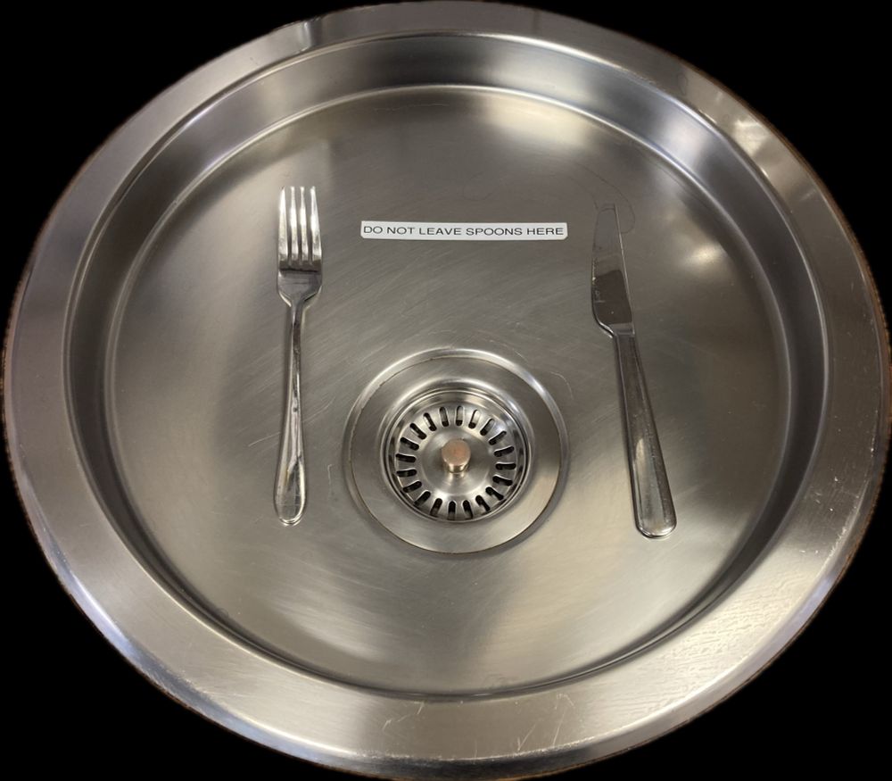 A kitchen sink with a label that says “DO NOT LEAVE SPOONS HERE”. Some miscreant has left a fork and knife there instead. 
