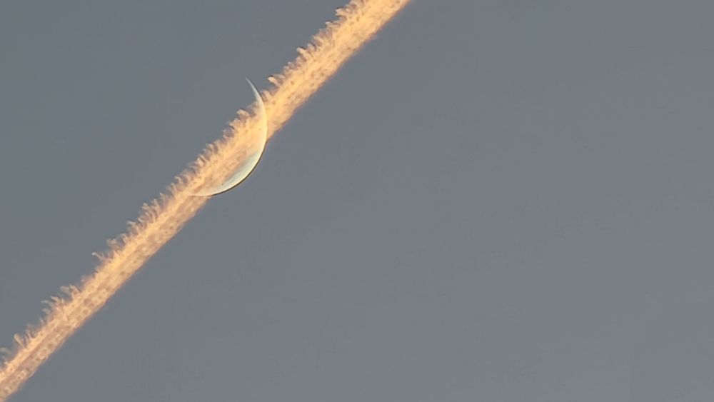 Contrail passing in front of crescent moon at sunset