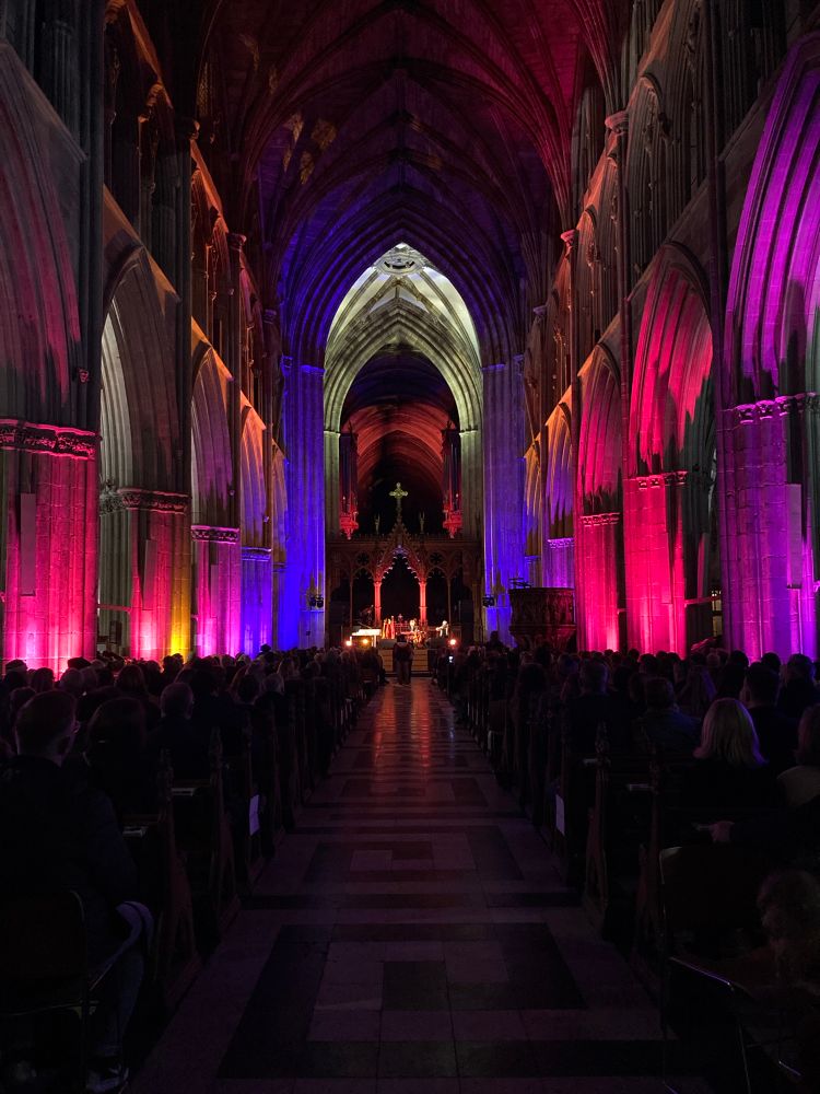Photo looking down the nave of a darkened cathedral lit in lovely magentas and blues. At the very end two women play keyboard and violin on the stage set up in front of the pulpit. 