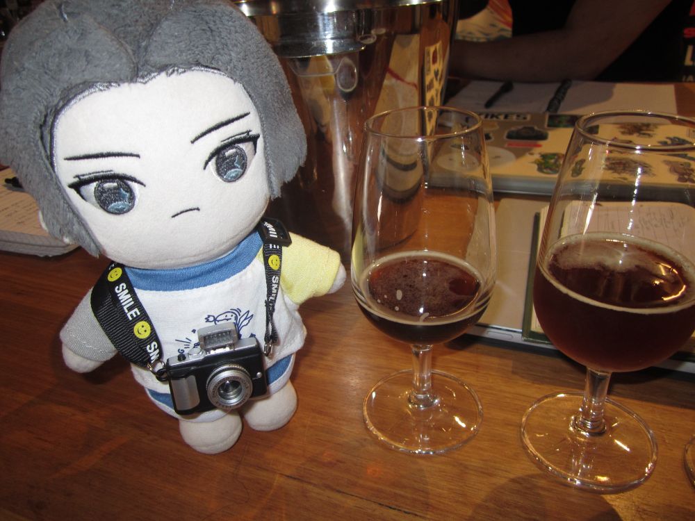 photo of jiang cheng in same outfit as above skeet. he's standing on a brown wooden table next to two tulip glasses of dark beer