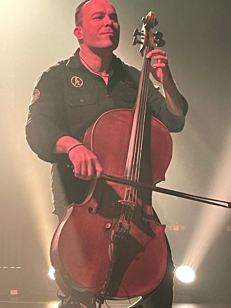 Photo of a man with short hair playing a cello and looking happy. The lighting is bright and white