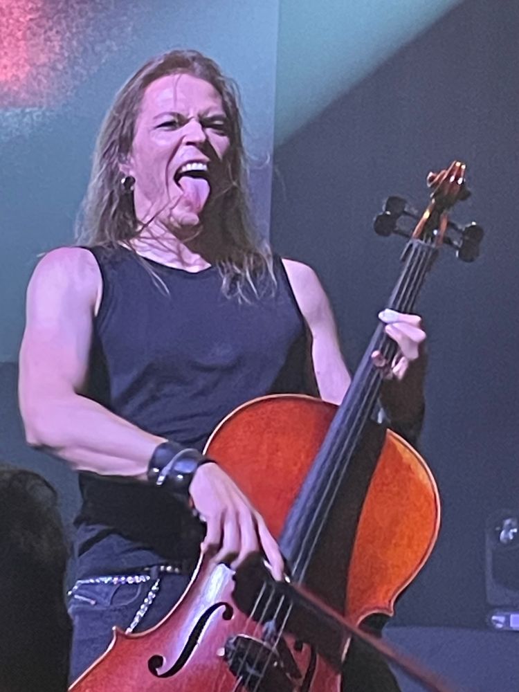Photo of a blond long haired man playing cello on stage and sticking his tongue out