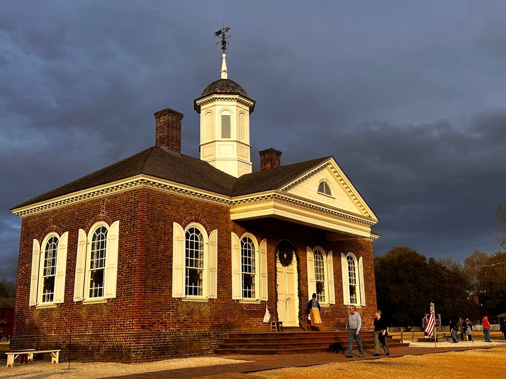 The Colonial Williamsburg, Virginia, USA courthouse warmly lit from a setting sun with dark storm clouds filling the sky in contrast. 