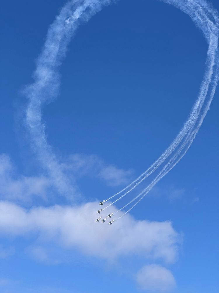 A formation six planes completing a loop with smoke against a blue sky. A few clouds in the background