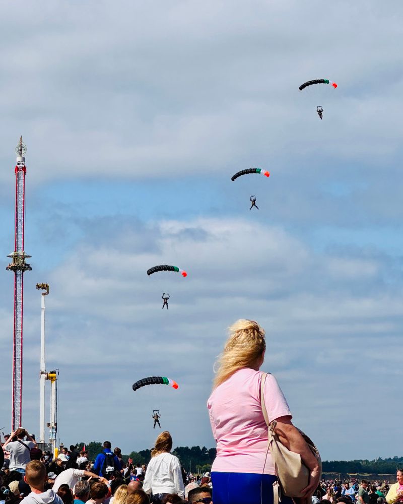 Four parachutists landing near the beach. A crowd of people and a woman in a pink top are in the foreground.