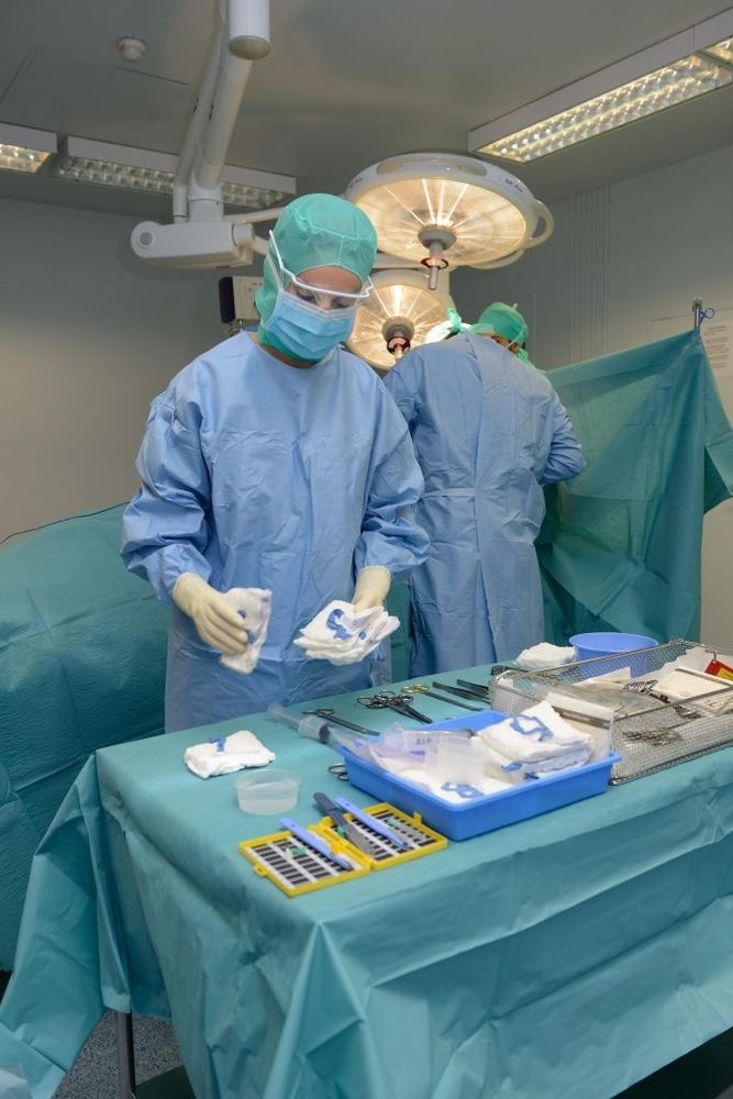 Un bloc opératoire. Au premier plan, une table (couverte d'une nappe stérile verte) sur laquelle sont en train d'être disposés les nombreux objets qui seront employés pendant la chirurgie: compresses, scalpels, ciseaux, seringues, etc. Derrière la table, une infirmière de bloc opératoire en tenue complète (charlotte, masque, blouse stérile jetable bleue, gants stériles) disposes ce qui semble être des compresses. Derrière elle, de dos, un infirmier de opératoire ou un chirurgien, dans la même tenue, se tient devant un champ opératoire stérile vert surplombé par des lampes de bloc opératoire. Bref, une femme aux cheveux couverts portant une robe ample couvrant tout son corps. Et devant des objets tranchants. C'est absolument terrifiant. (Non. Les gens qui ont édicté la procédure décrite dans l'article que je qrt sont juste des gros racelards de merde, collabos zélés et enthousiastes d'un régime raciste et islamophobe)