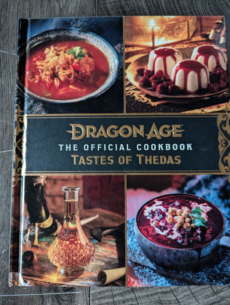 Hardback book with pictures (small images, I think one is a beet soup, there's a jar of amber liquid, and some cream dessert with a red sauce of some kind) of different foods on the cover.

Text says, "Dragon Age: Tastes of Thedas. The official cookbook."