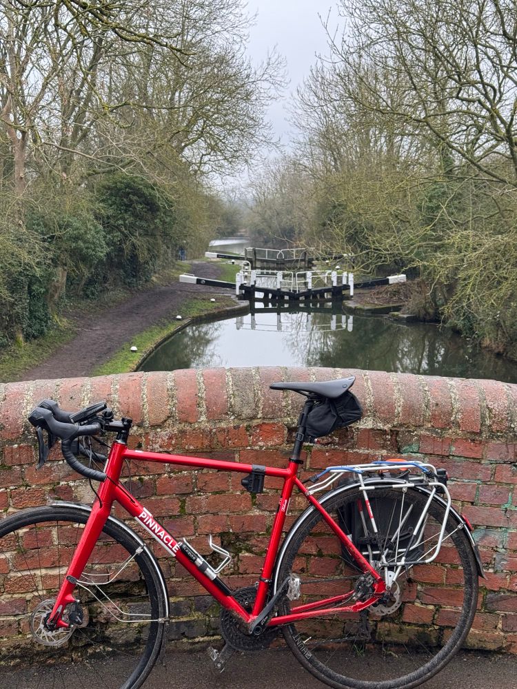A red drop bar bicycle with rack and pannier leans against the old brick wall of a hump-backed bridge over a canal with a lock gate seen in the distance