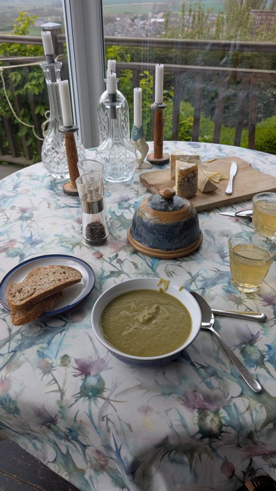 A bowl of rather thick green soup on a table with brown bread, a butter dish and some glasses of homemade kombucha. Behind them are some candles and a chopping board with cheese on it. There is a glimpse of garden beyond.
