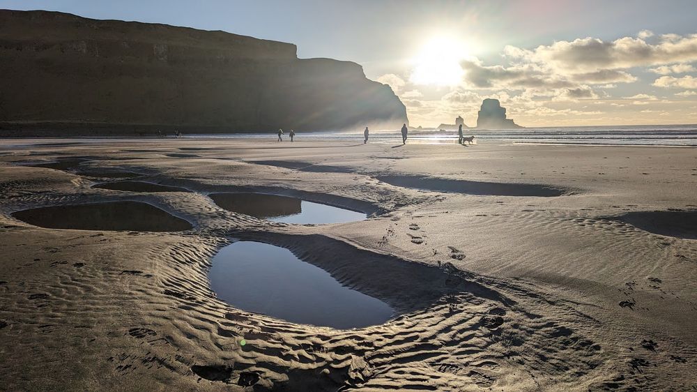 Talisker beach on the Isle of Skye looking into the sun with striking pools of water in the foreground and tiny silhouetted figures in the distance.