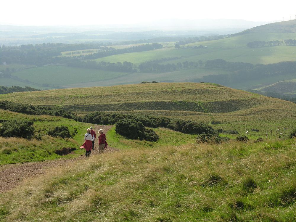 The intriguing Castlelaw Hill Fort, high on the eastern flank of Castlelaw Hill in the Pentland Hills south of Edinburgh. The image shows a path with three people on it leading down to the hillfort, with its ramparts visible in the background. The scene is in sunlight though there is haze in the distance.