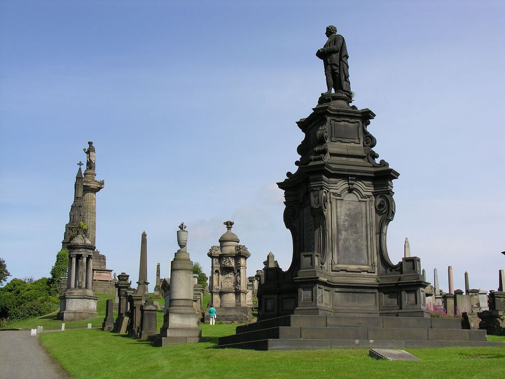 Glasgow's amazing Necropolis. The image shows a view up a hill of mown grass with a path in the lower left of the frame. The hillside is covered by extremely large memorials in mid or dark grey. The scale is set by a person in the bottom centre of the frame. The sky is blue.