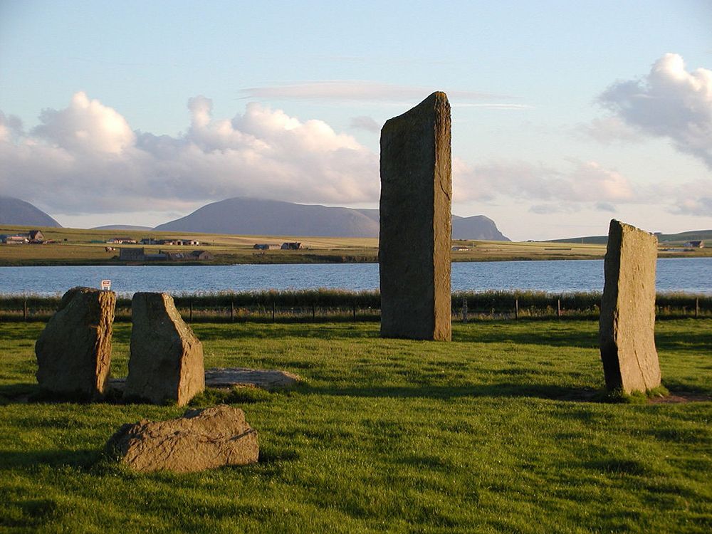 The Stones of Stenness circle and henge on Orkney's West Mainland. The image shows a grassy area in the foreground with a road behind then a loch cutting across the frame and low-lying land behind it. In the distance are hills. There are four standing stones in the grassy area, two low ones on the left, a taller one on the right, and the tallest in the centre of the frame. Another stone is lying on the ground in the left foreground. Sunlight is coming from the right.