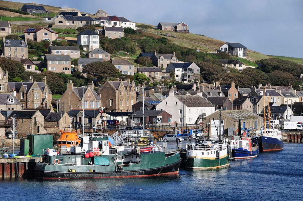 There's nowhere quite like Stromness in Orkney. The image shows Stromness built on rising ground beyond its harbour, with fishing boats moored in the foreground. The buildings are mainly of stone or finished in white and the scene is in sunlight.