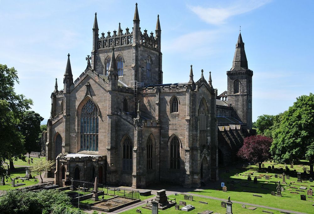 Dunfermline Abbey Church. The image shows a view of the church from a perspective level with its upper parts and from a corner. The larger part of the church is slightly to the left of the centre of the frame and the rest of it tails off to the right and behind. The church is surrounded by a graveyard and by trees in green leaf. The scene is in sunlight.