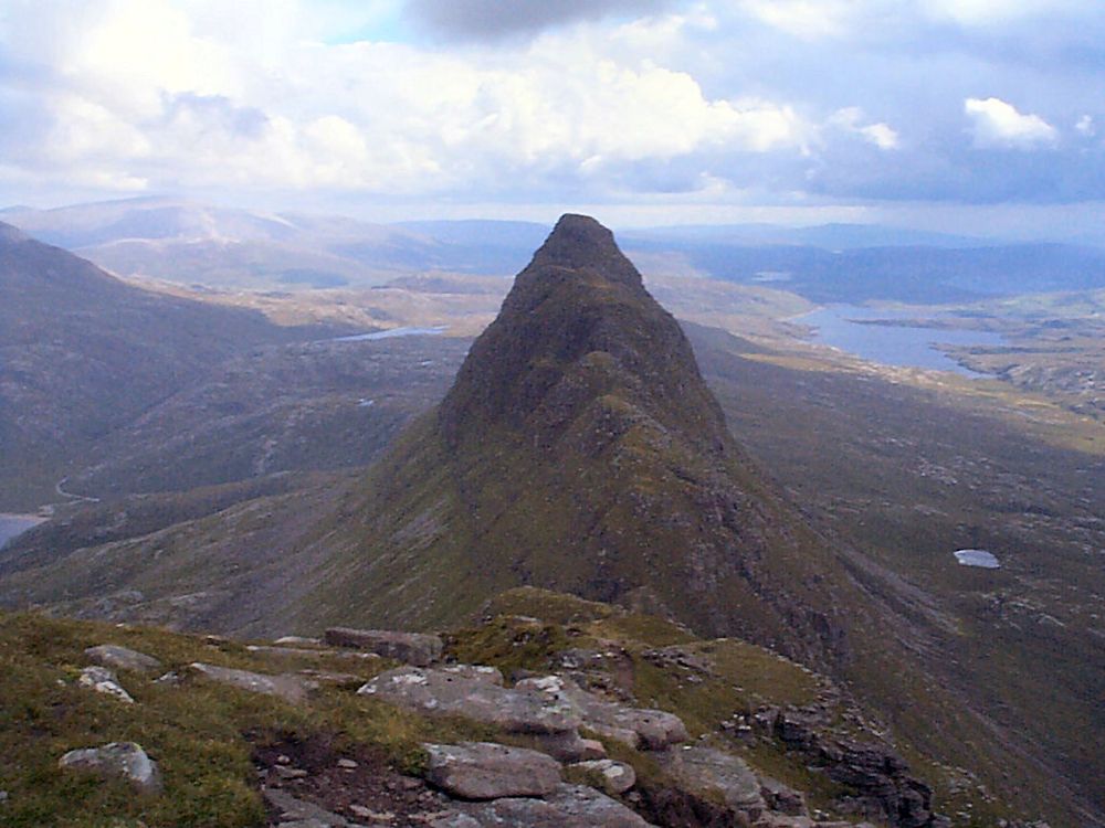The view east from the top of Suilven, inland from Lochinver on the west coast of Sutherland. The image shows descending rocky ground in the foreground leading to a narrow neck and then climbing to a sharply pointing peak, which is slightly lower than us. In the background is much lower lying land of stony ground with rivers and lochs. The sunlight is patchy.