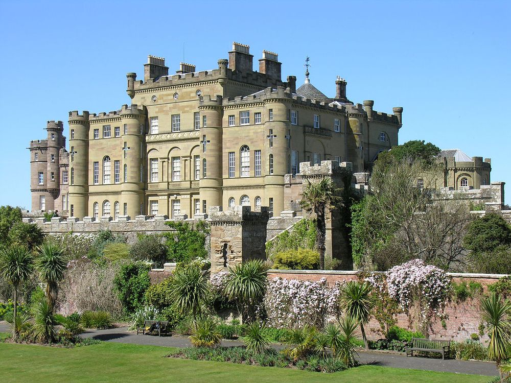 Culzean Castle. The image shows a view across an ornate sunken garden to a very grand castle built in honey-coloured stone with its long face pointing out to the left of the frame. It has several different blocks rising to the highest in the centre of the structure and there are many large windows. The sky is blue.