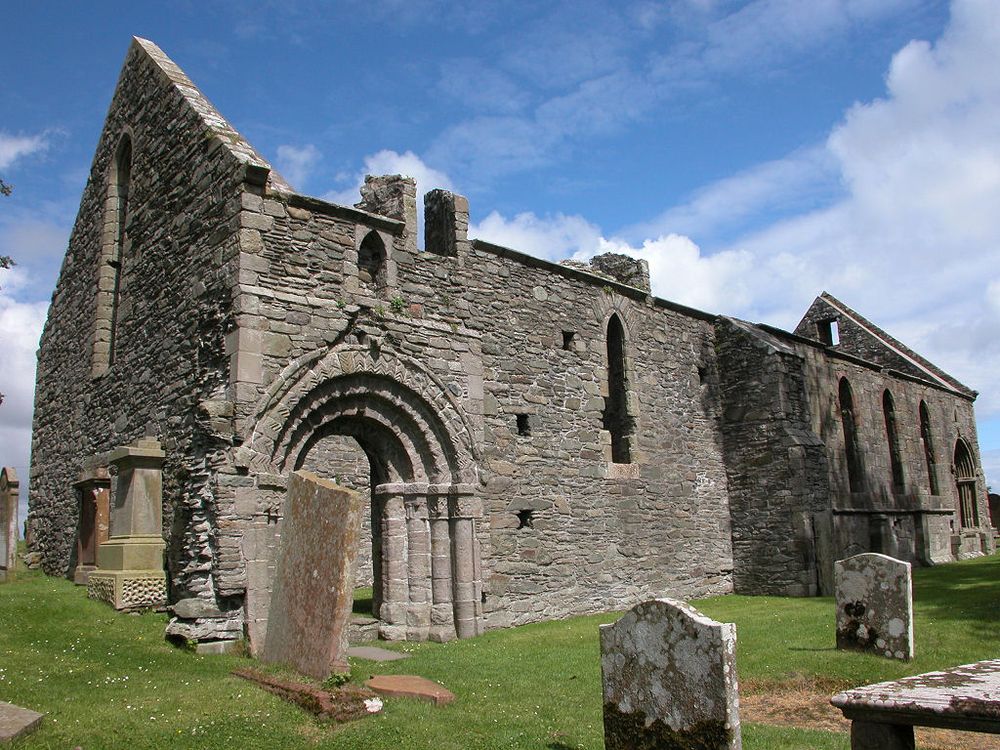 The image shows a roofless grey stone church from an oblique angle from the left centre of the frame back into it on the right. There is an ornate arched doorway on the side of the near corner and tall window openings along the church. There are gravestones in the foreground. The scene is in sunlight.