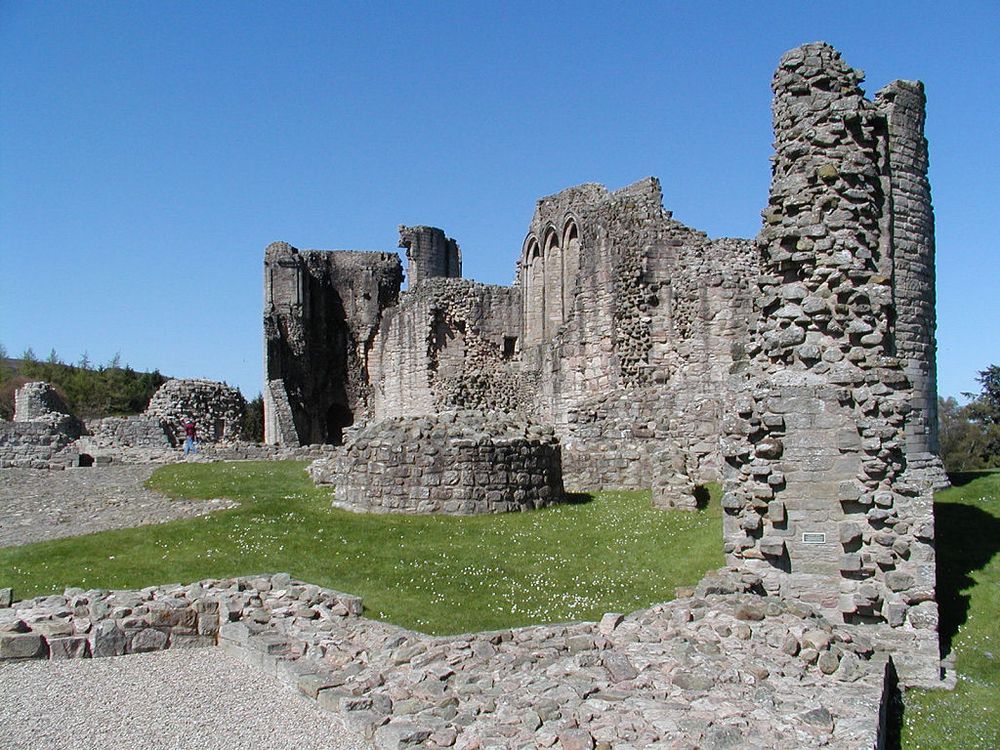 Kildrummy Castle in Aberdeenshire. The image shows the grey stone ruins of the castle under a clear blue sky. A wall on the right of the frame extends to considerable height and continues round to the middle of the frame. The bases of other stone ruins can be seen, including in the foreground. Areas of grass have daisies growing.