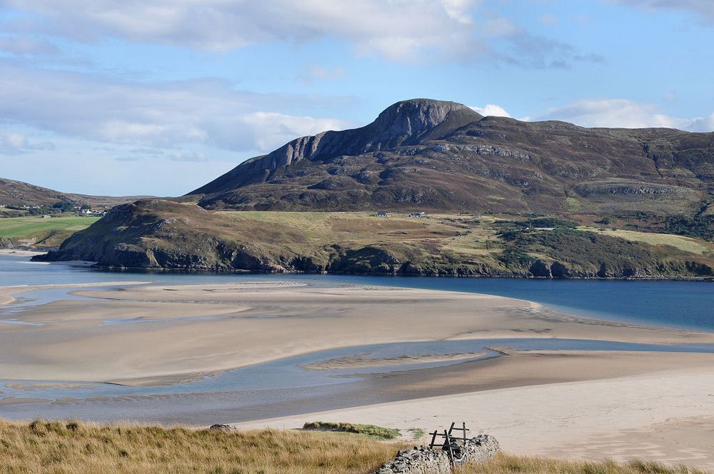 The view across Tongue Bay from the road to Talmine on the north coast of Sutherland. The image shows a view down a rough grassy slope to a broad inlet from the sea that is largely covered by sandbanks, with a blue channel on the far side. This is below the far shore, which climbs to a rocky hill. The scene is in sunlight.