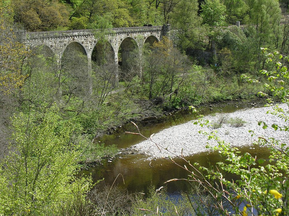 Killiecrankie in Perthshire. The image shows a view from a high viewpoint down through trees into a river valley with a river whose brown water divides past a stone bank in its centre. In the background a path can be seen beyond the river bank and above it is a railway viaduct and embankment. 