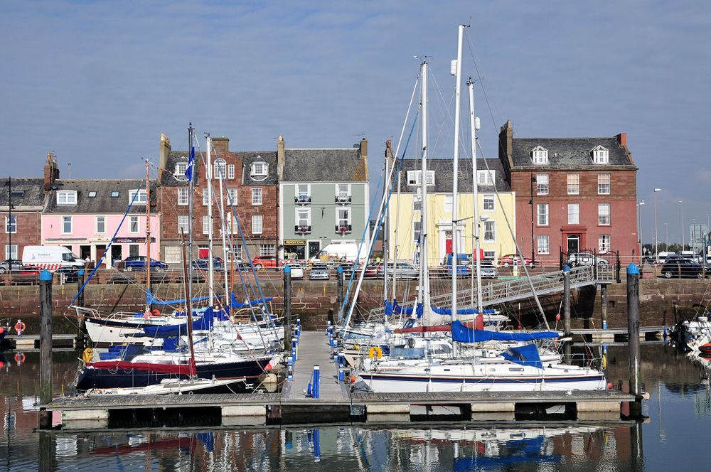 Arbroath in Angus. The image shows a view across a harbour. The lower centre of the frame is occupied by a line of pontoons stretching away from the viewer, at which large yachts are moored on both sides. Then there is a harbour wall and above it are cars parked on a road with a line of mainly three-storey buildings on the road’s far side, made of brick or with pastel finishes. The sky is blue.