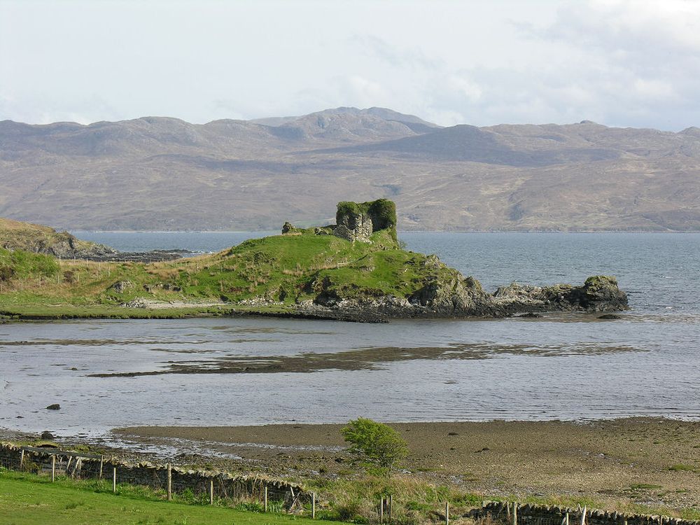 The remains of Knock Castle on the Isle of Skye. The image shows a view down onto a shorline with a rocky headland protruding into it from the left in the middle distance. On the highest point of the headland are stone ruins covered in vegetation. The sea is in the background with, beyond that, mountains on a far shoreline.